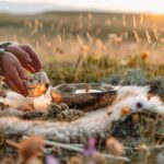 Hands lighting a candle beside a ceremonial bowl and natural elements in an outdoor ritual setting at sunset