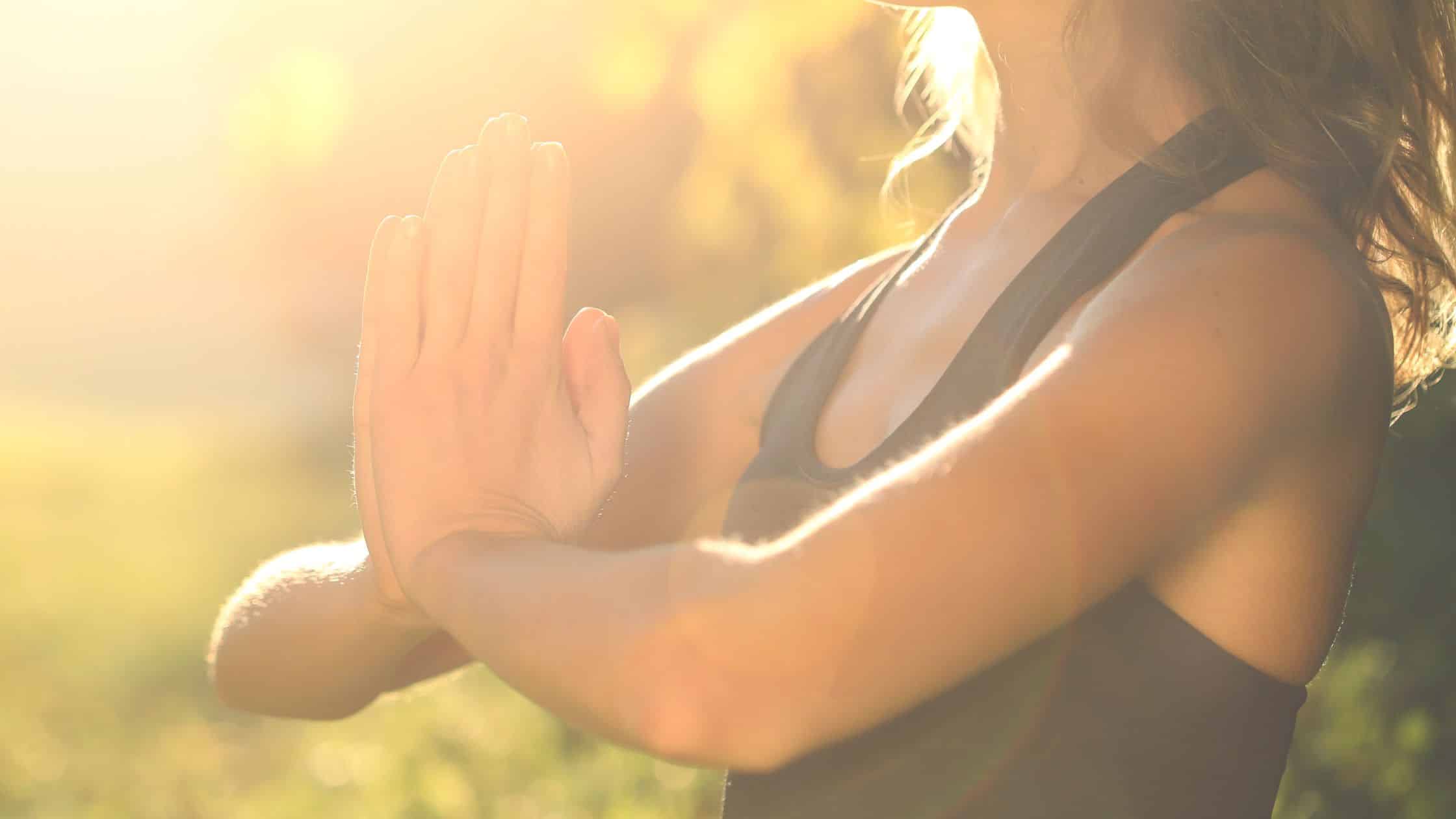 A woman practicing meditation outdoors in warm sunlight, hands in prayer position, symbolizing trust, clarity, and spiritual alignment.